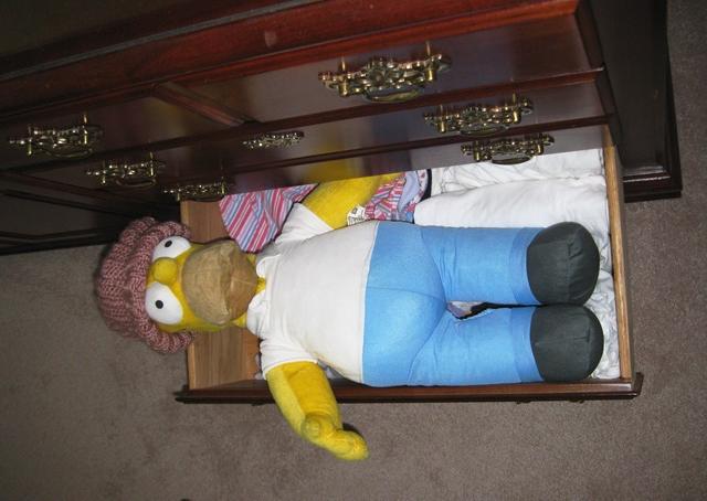 Homer Simpson doll is reclining in the dresser drawer wearing a hat, knitted in Lynda Lambert’s pattern by Donna W. Hill, using Caron’s Simply Soft Victorian Rose yarn; women’s undies visible : Homer, courtesy of Mary McAleavey; photo by Rich Hill.